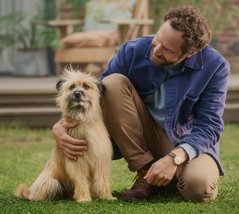 Ein Mann kniet auf dem Rasen und hat eine Hand um einen Hund gelegt, den er anschaut.